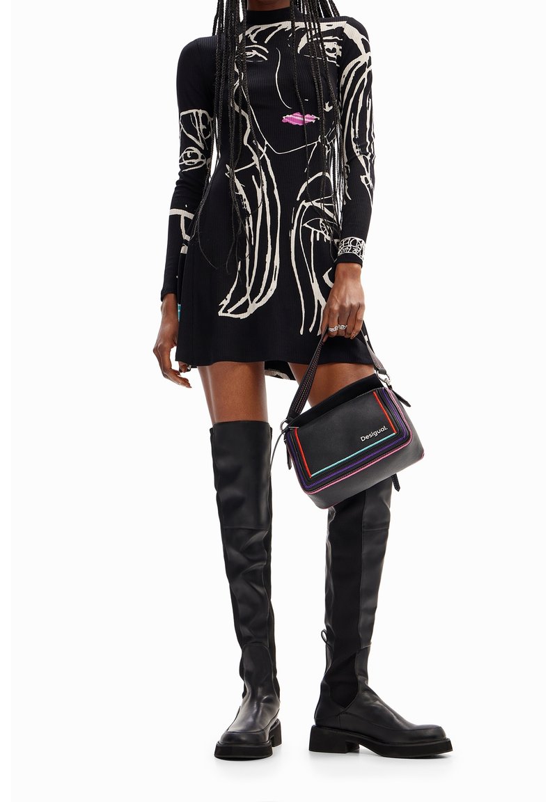 Femme portant une robe noire à manches longues avec un motif abstrait de visage blanc, des bottes noires montant au-dessus des genoux, et tenant un sac à main noir à garniture colorée.