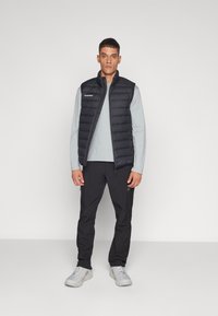 Zwarte gewatteerde vest over een lichtgrijs longsl sleeve shirt, gecombineerd met zwarte broek en lichtgrijze schoenen. Het vest heeft horizontale stiksels.