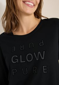 Czarny top z długim rękawem i okrągłym dekoltem, z teksturowanym czarnym napisem "GLOW PURE" umieszczonym w pionie i poziomie.