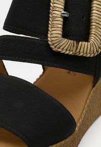 Μαύρο πλατφόρμα πέδιλο με δύο λουριά, υφή ψεύτικου δέρματος και μεγάλη υφαντή αγκράφα. Η πλατφόρμα έχει φυσικό σχέδιο σχοινιού.