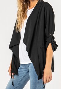 Femme portant un cardigan noir ouvert sur le devant avec les manches retroussées, un t-shirt blanc et un jean bleu clair, debout devant un fond uni.