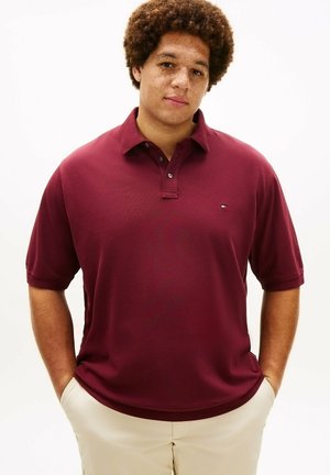 Ung mand med krøllet hår og fregner iført en bordeaux polo og beige bukser med hænderne i lommerne, stående mod en hvid baggrund.