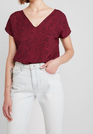 Woman wearing a red, short-sleeve, V-neck patterned blouse tucked into high-waisted white jeans, with hand in pocket.