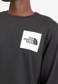 Černé tričko s dlouhým rukávem vyrobené z bavlny, s bílou čtvercovou nášivkou s logem "THE NORTH FACE" v tučném černém písmu.