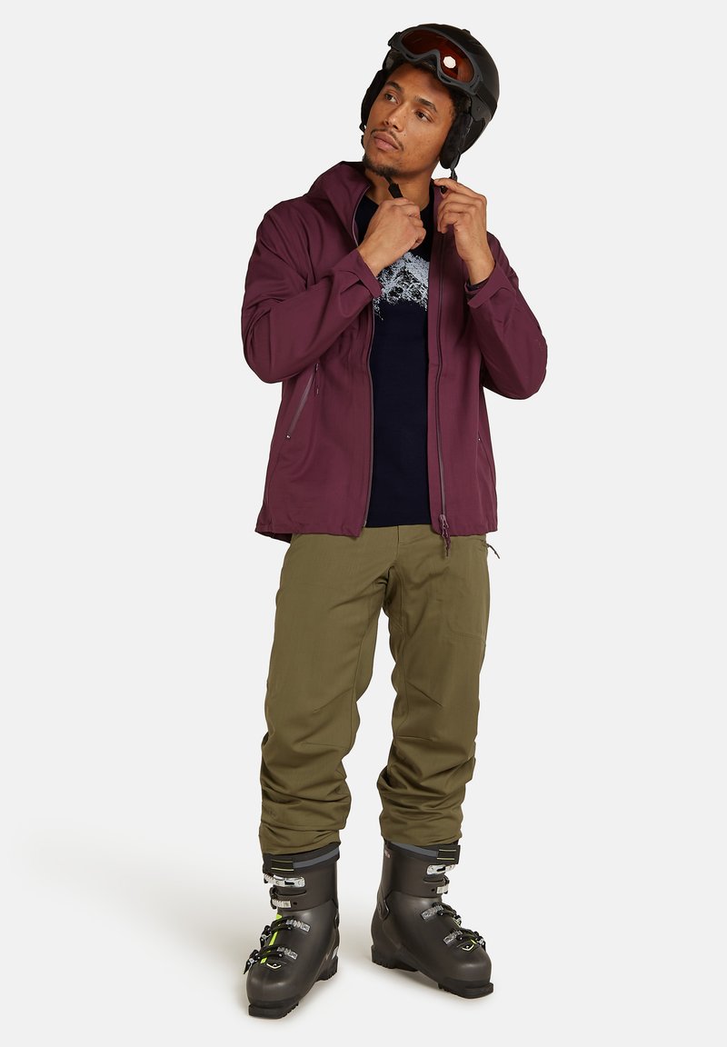 Skidkläder som inkluderar en vinröd jacka med blixtlåsfickor, en marinblå tröja med grafisk design, olivgröna byxor och svarta skidstövlar.