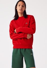 Sweatshirt rouge avec col rond et poignets côtelés, associé à un short vert avec une bande latérale blanche et un logo en accent.