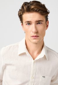 Νέος άνδρας με ανοιχτό καφέ μαλλιά και μπλε μάτια, φορώντας ένα λευκό πουκάμισο με κουμπιά και το λογότυπο Wrangler στην τσέπη, με ουδέτερο φόντο.