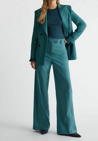 Frau inantalon aus glänzendem Material in Teal, passendem Blazer und glitzerndem, geripptem Oberteil, die mit einer Hand auf der Hüfte vor einem weißen Hintergrund steht.