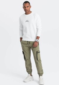Sweatshirt blanc avec logo noir, pantalon cargo vert olive avec zips latéraux et baskets blanches. Le mannequin se tient les mains dans les poches.