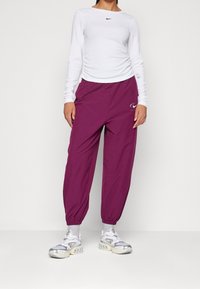 Kvinna i vit långärmad topp och lila joggingbyxor med elastiska muddar, kombinerat med vita och silvriga träningsskor mot en neutral bakgrund.