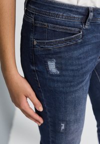 Jeans de denim azul oscuro con corte ajustado, que presentan un parche desgastado y múltiples bolsillos. Detalle de costura visible en las costuras.