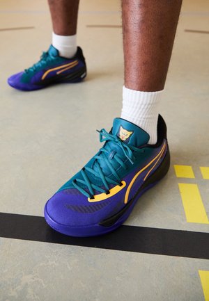 Zapatillas deportivas en púrpura y verde azulado con detalles en negro, parte superior de textil, cordones vibrantes y logo en la lengüeta, situadas sobre el suelo de un gimnasio.