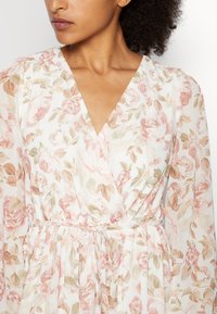 Vestido floral de tela ligera con escote en V, mangas largas abullonadas y cintura con lazo, con un patrón de flores en tonos pastel rosa y crema.