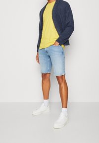 Cárdigan de punto azul con botones, camiseta amarilla, pantalones cortos de mezclilla azul claro y zapatillas blancas con calcetines al tobillo. Texturas suaves y corte casual.