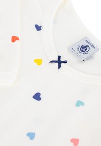 Λευκό βαμβακερό τοπ με κοντά μανίκια που διαθέτει μοτίβο με καρδιές σε κόκκινο, κίτρινο, μπλε και ναυτικό μπλε, με λεπτομέρεια φιόγκου στο ναυτικό μπλε στο λαιμό.