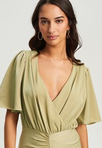 Olivgrünes Wickelkleid mit kurzen, ausgestelltenÄrmeln, tiefem V-Ausschnitt und gerafftem Taillendetail, aus weichem, fließendem Stoff. Goldene Creolen.