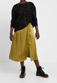 Maglione nero scintillante con scollo rotondo, abbinato a una gonna in raso dorato con rifinitura in pizzo e spacco frontale. Stivaletti neri con lacci.