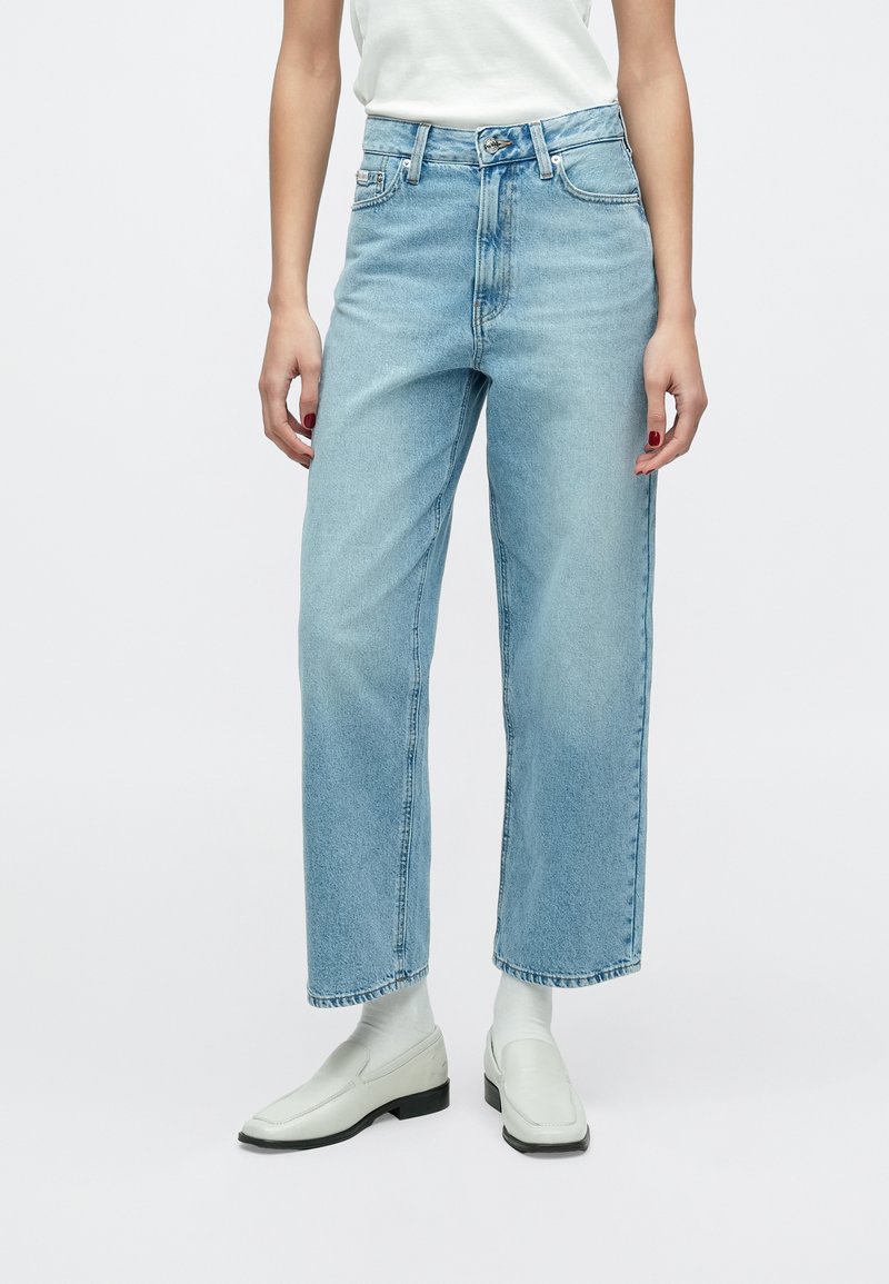 Világoskék farmernadrág, bő szárú, magas derekú, finoman kopott részletekkel, egyszerű fehér cipővel párosítva.