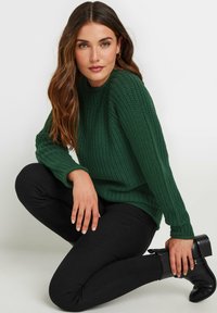 Groene gebreide trui met een gestructureerd patroon, ronde halslijn en geribde boorden, gecombineerd met zwarte slim-fit broek en zwarte enkellaarsjes.