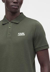 Homme portant un polo vert olive à trois boutons avec le logo blanc "KARL LAGERFELD" sur la poitrine gauche sur un fond uni.