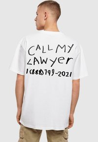Hvid oversized t-shirt med fed sort tekst på ryggen, der står "KALD MIN LÆGER" med et telefonnummer nedenunder. Korte ærmer, rund hals.