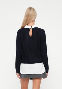 Marineblauwe gebreide trui met lange mouwen, voorzien van een keyhole-detail op de rug en een zachte textuur, gelaagd over een wit shirt en een grijze rok.