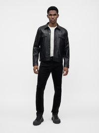 Jeune homme debout portant une veste en cuir noire, un t-shirt blanc, un pantalon noir et des baskets noires sur un fond uni.