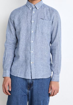Homme portant une chemise à manches longues à carreaux bleu clair avec une poche poitrine et un jean bleu sur un fond uni.