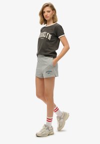 Pantalones cortos de algodón grises con bolsillos laterales y un logo azul de "SUPERDRY NYC". Combinados con una camiseta gris oscura que presenta "BROOKLYN" en blanco.