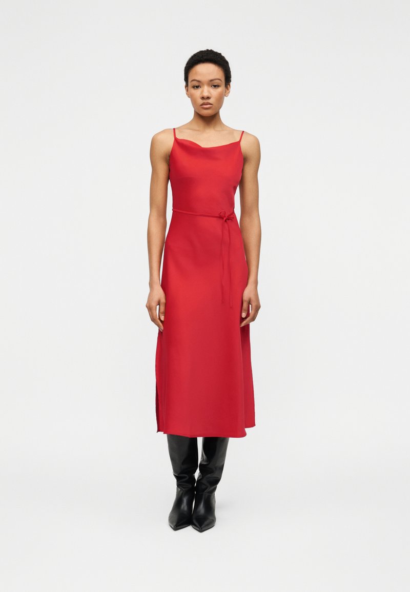 Γυναίκα που στέκεται φορώντας κόκκινο αμάνικο μεσαίο φόρεμα με πλευρική σχισμή και μαύρες μπότες μέχρι το γόνατο με μυτερό άκρο, κοιτάζοντας εμπρός.