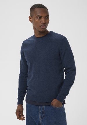 Homme portant un pull bleu marine et un jean, debout avec une main dans la poche, regardant sur le côté contre un fond clair uni.