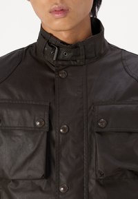 Μαύρο, υφασμάτινο σακάκι με ψηλό γιακά, κουμπιά με κλιπ και δύο μπροστινές τσέπες. Διαθέτει λεία επιφάνεια και ελάχιστες ραφές.