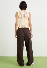 Pletená háčkovaná tílko v krémové barvě s květinovým vzorem, spárované s volnými tmavohnědými kalhotami a šedými teniskami na zelené podlaze.