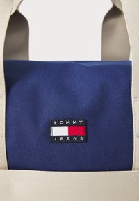 Námořnická plátěná taška s béžovými popruhy, na které je obdélníková černá nášivka s nápisem "TOMMY JEANS" a červenobílo-modrou grafikou.