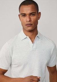 Světle šedé polo tričko vyrobené z měkké tkaniny má tříknoflíkovou légu a malé logo na levém hrudníku. Krátké rukávy a volný střih.