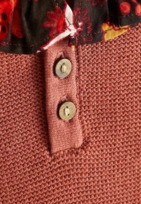 Tissu en maille couleur rouille avec deux boutons marron sur une patte de boutonnage, sous un col en tissu noir et rouge à motifs floraux.