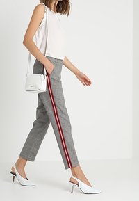 Un pantalon à carreaux gris avec une bande latérale rouge, associé à un haut sans manches blanc et des escarpins blancs à talons. Un petit sac à bandoulière blanc est inclus.