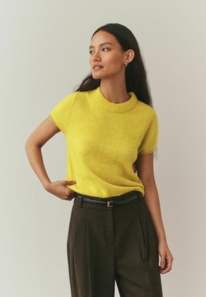 Short-sleeve yellow sweater with a ribbed neckline, paired with brown high-waisted trousers and a black belt. Smooth texture, casual fit.
