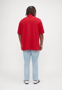 Rød kortærmet polo shirt med afslappet pasform, parret med lyseblå jeans og sorte og hvide sneakers, set fra ryggen.