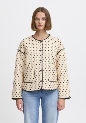 Vrouw in een beige jas met lange mouwen met zwarte stippen, zwarte rand, zakken aan de voorkant en blauwe spijkerbroek, staand tegen een effen witte achtergrond.