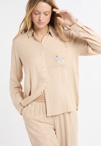 Beżowy, pasiasty zestaw piżamowy z długim rękawem, zapinany na guziki, z kieszenią na piersi. Kieszeń ozdobiona grafiką psa. Miękka tkanina.