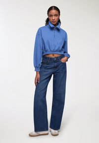 Felpa blu corta con colletto zip, abbinata a jeans larghi blu scuro e sneakers bianche con dettagli beige.