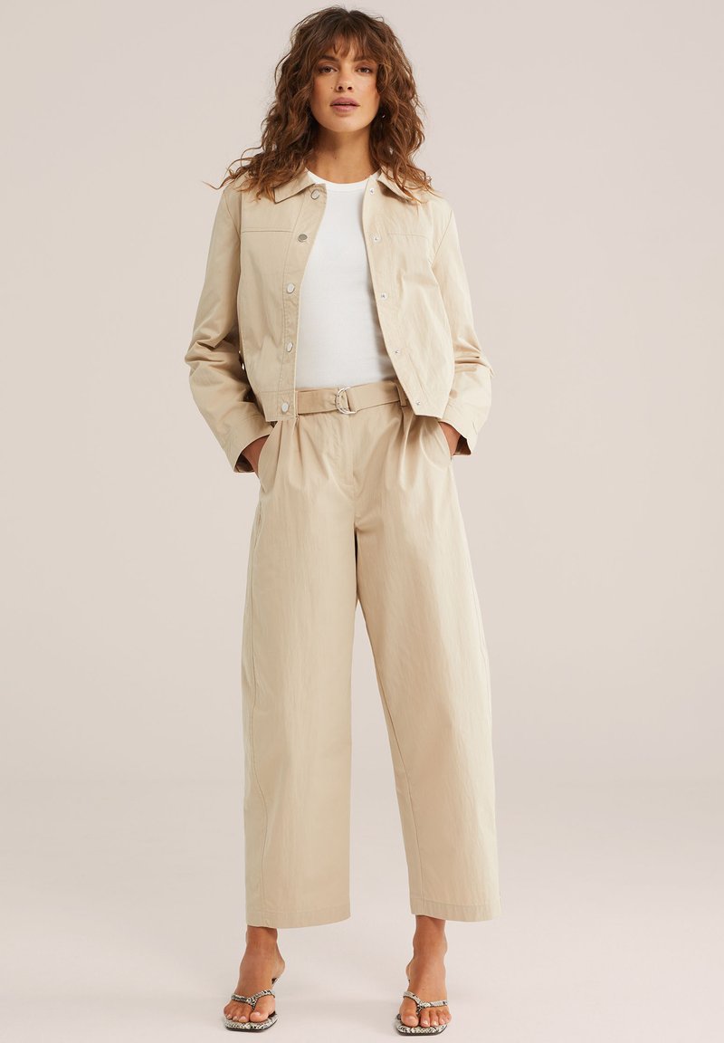 Femme portant une veste courte beige et un pantalon large taille haute avec un haut blanc et des sandales à talons, debout les mains dans les poches.