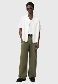 Chemise blanche à manches courtes, pantalon large vert olive, chaussures noires, collier en chaîne argenté et lunettes de soleil rectangulaires. Design simple, coupe décontractée.