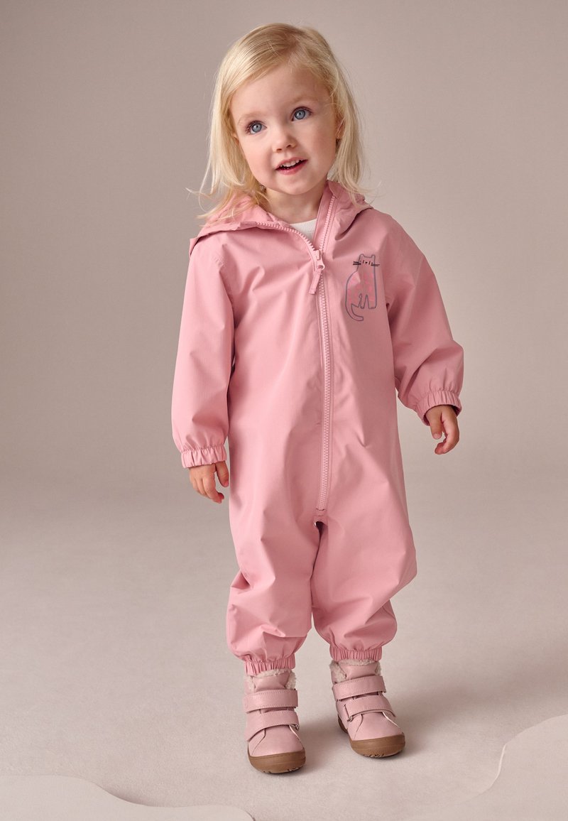 Bambino piccolo con capelli biondi che indossa una tuta impermeabile rosa con cappuccio decorata con un motivo a gatto e stivali rosa, in piedi su un pavimento chiaro contro uno sfondo neutro.