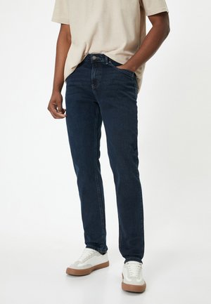 Dunkelblaue Denim-Jeans mit schlankem Schnitt, ausgestattet mit einem Knopfverschluss, fünf Taschen und minimalen Nähdetails. Getragen mit weißen Turnschuhen.