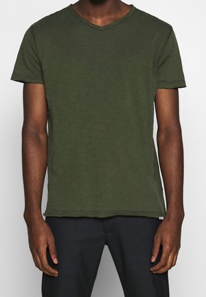 Mies, jolla on oliivinvihreä lyhythihainen V-kaula-aukollinen t-paita ja tummat housut, seisoo rennosti käsivarret sivuilla tasaisella taustalla.