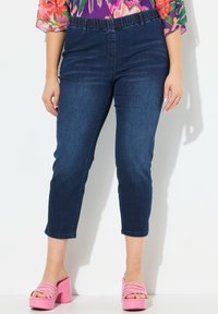 Ciemnoniebieskie dżinsowe spodnie capri z elastycznym pasem, o gładkiej fakturze i dopasowanym kroju. Noszone z różowymi sandałami na słupku.