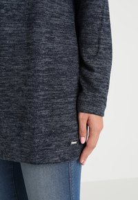 Marinblå tröja med strukturstickning, med långa ärmar, en avslappnad passform och ett litet metalliskt logomärke vid nederkanten.