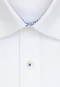 Chemise blanche en tissu texturé, couture bleue à l'intérieur du col et un bouton blanc avec un fil bleu au centre devant.